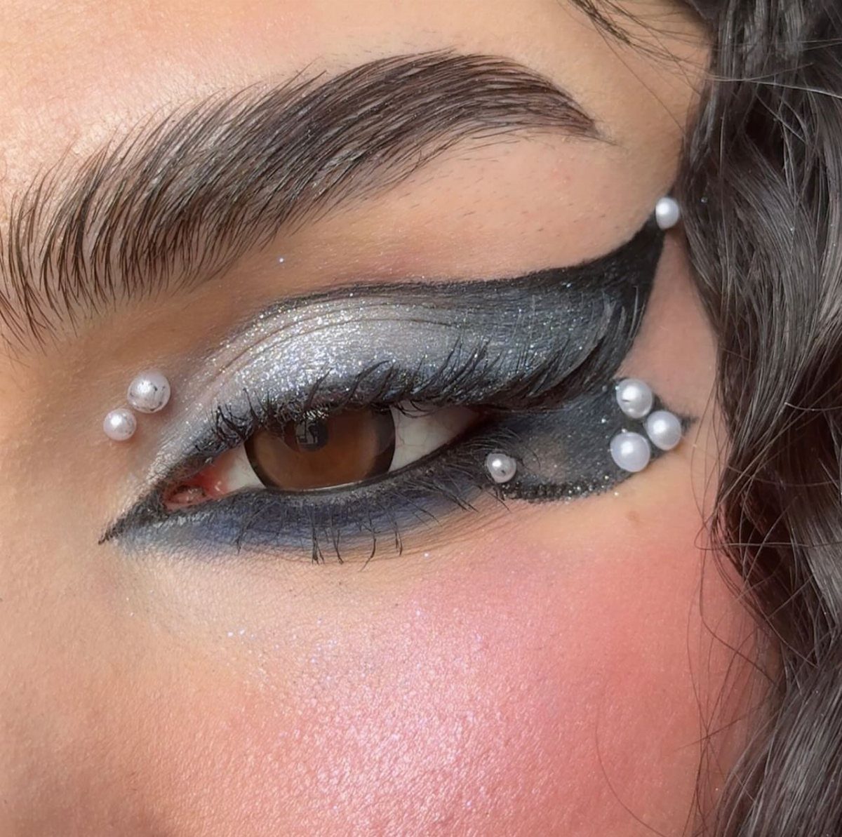 Eye with silver glitter eyeshadow, black winged liner, and decorative white pearls.