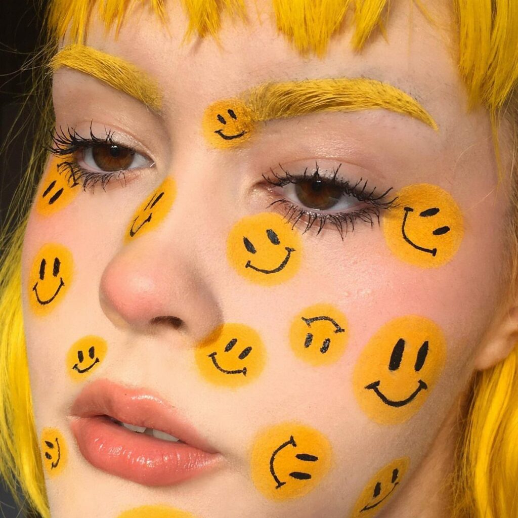Person with yellow hair and smiley face emojis painted on their face.