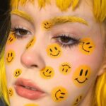 Person with yellow hair and smiley face emojis painted on their face.