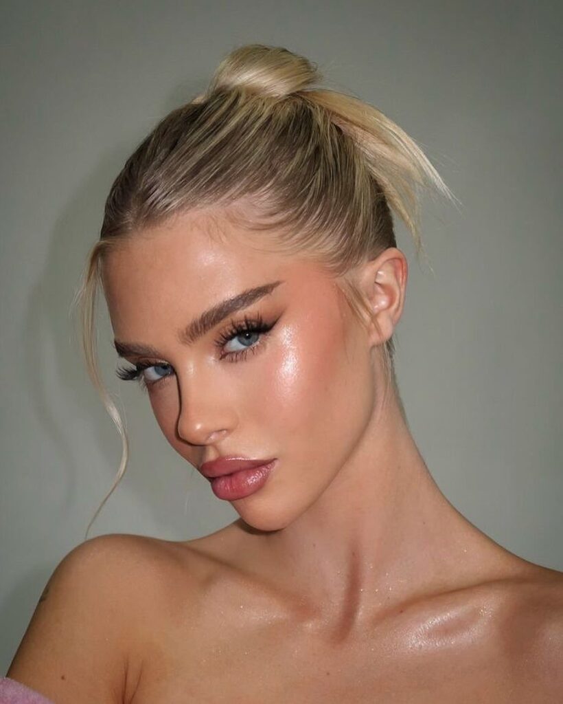 Woman with luminous, glowy skin makeup, glossy lips, and blonde hair in a bun.