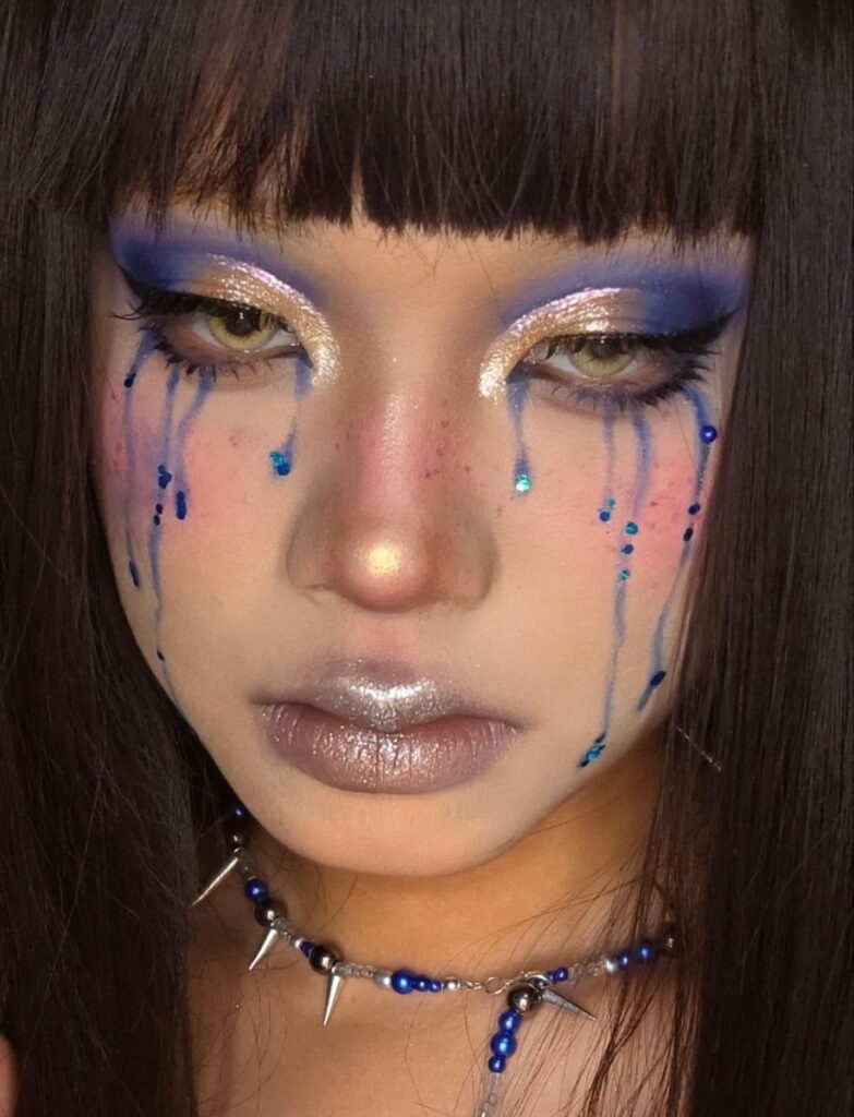 Person with blue and gold makeup and blue rain-like droplets on their cheeks.