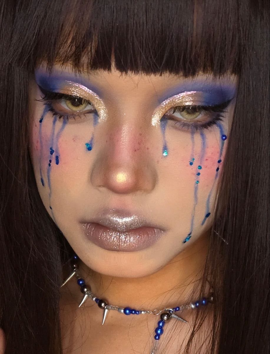 Person with blue and gold makeup and blue rain-like droplets on their cheeks.