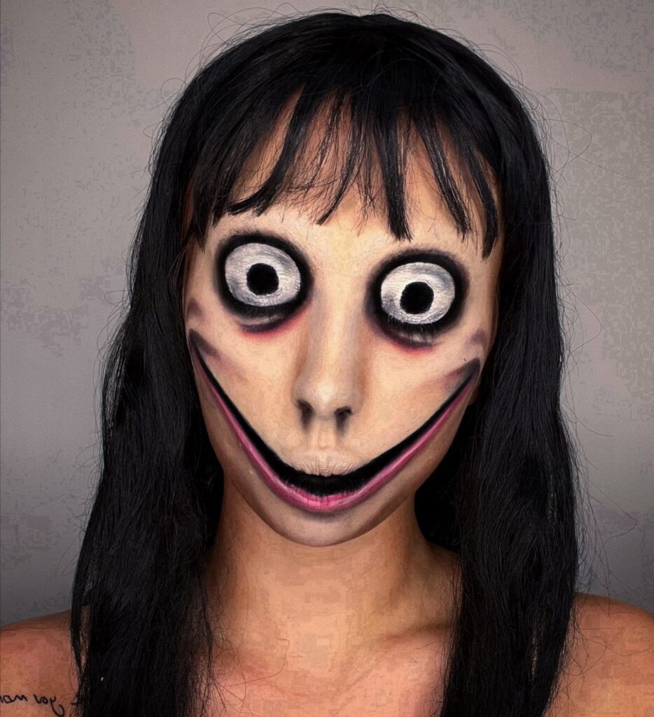 Person with long black hair wearing scary face paint with oversized eyes and wide smile