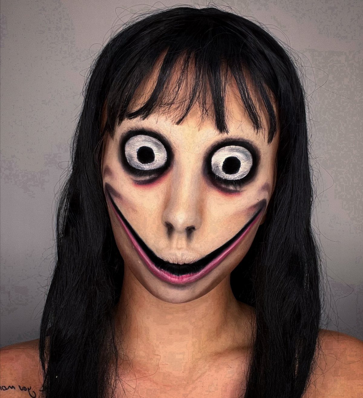 Person with long black hair wearing scary face paint with oversized eyes and wide smile