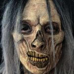 Zombie mask with pale wrinkled skin, skeletal nose, and yellowed teeth with grey hair.